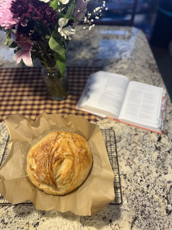 A golden crusty loaf cooling on parchment, next to an open book and a jar of fresh flowers